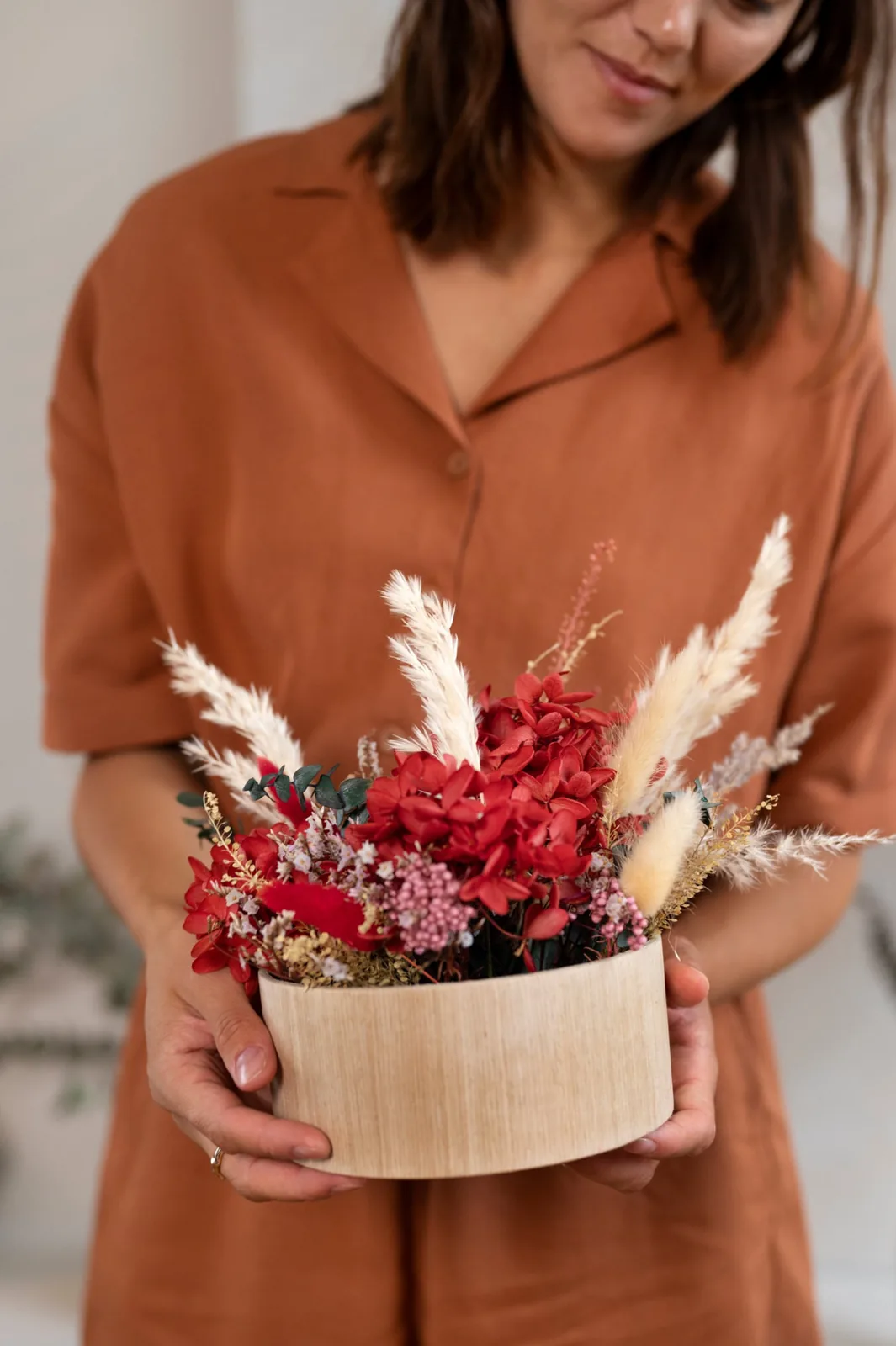 Donna che tiene tra le mani una composizione di ortensie rosse stabilizzate, eucalipto e pampas in un vaso di legno